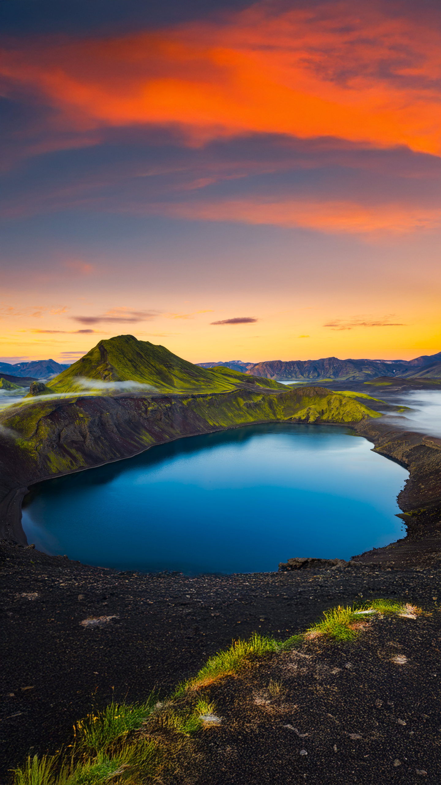 A serene volcanic lake nestles within a rugged, moss-covered landscape at sunset.