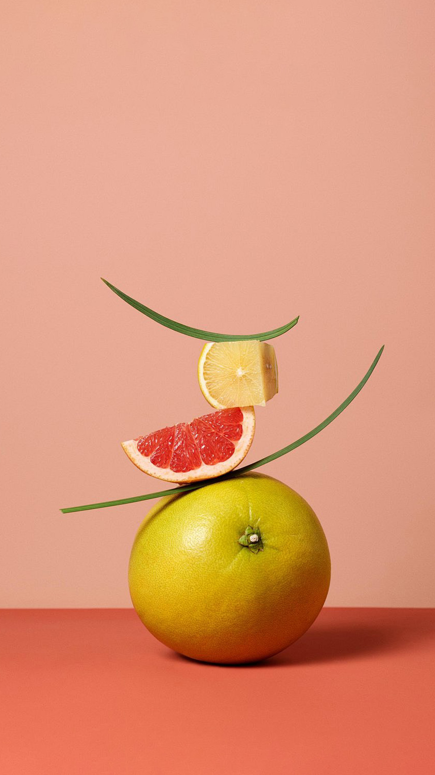 A vibrant yellow grapefruit is balanced on top of a red grapefruit, with a lemon slice and a green leaf curving around it.