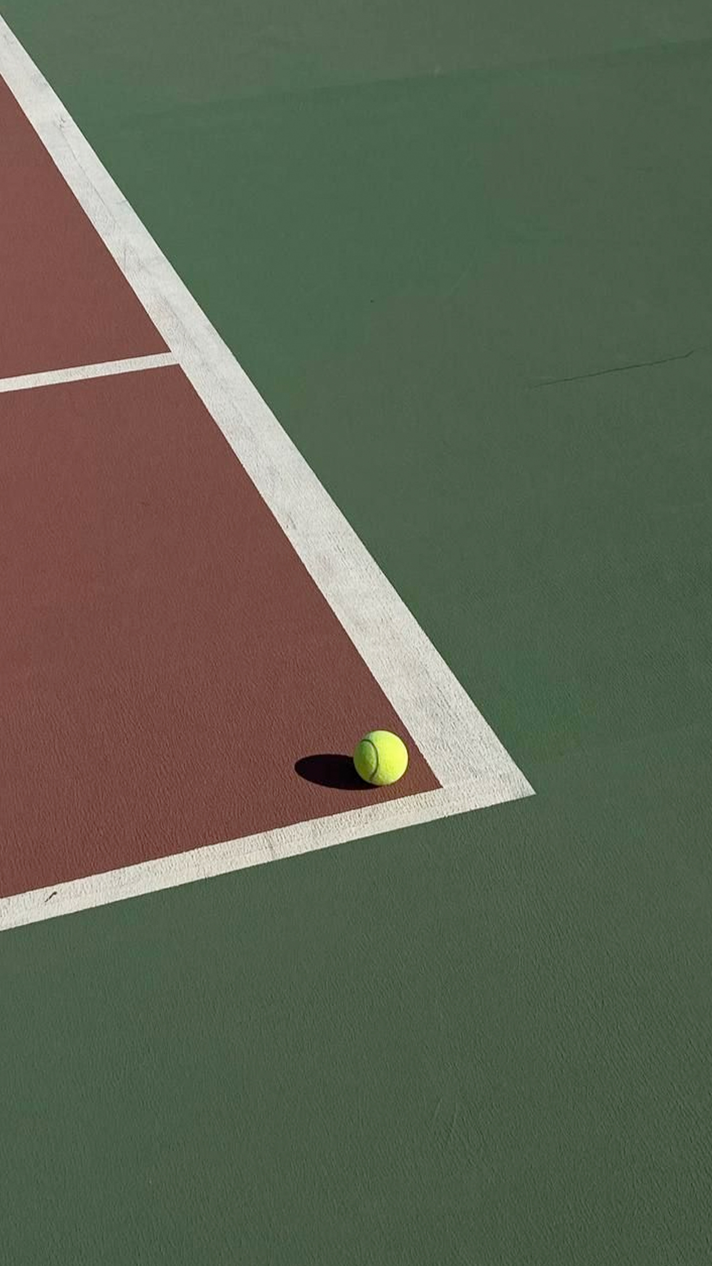 A tennis ball lies on the edge of a tennis court, with its shadow cast onto the surface.