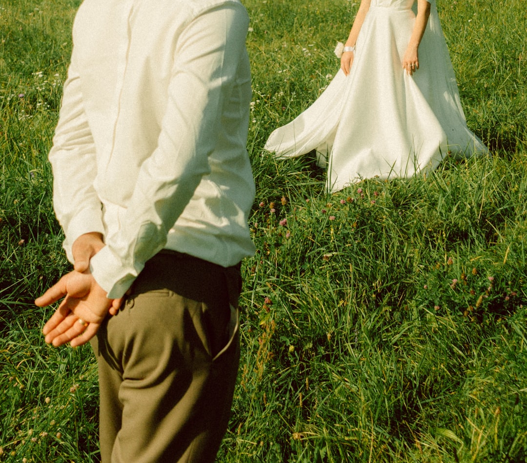A man stands with his back to the camera, his hands clasped behind him, while a woman in a white wedding dress walks through a lush green field.