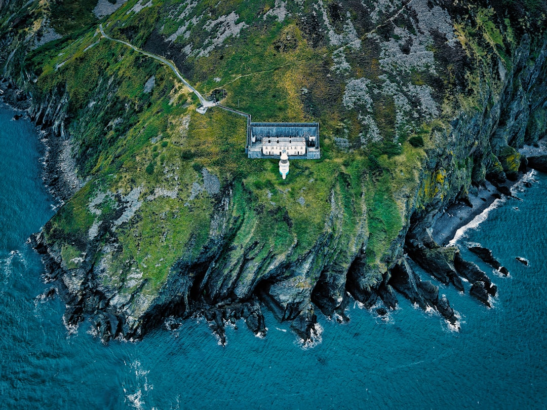 A rugged coastline with a white lighthouse and a building on a grassy cliff, surrounded by deep blue water.