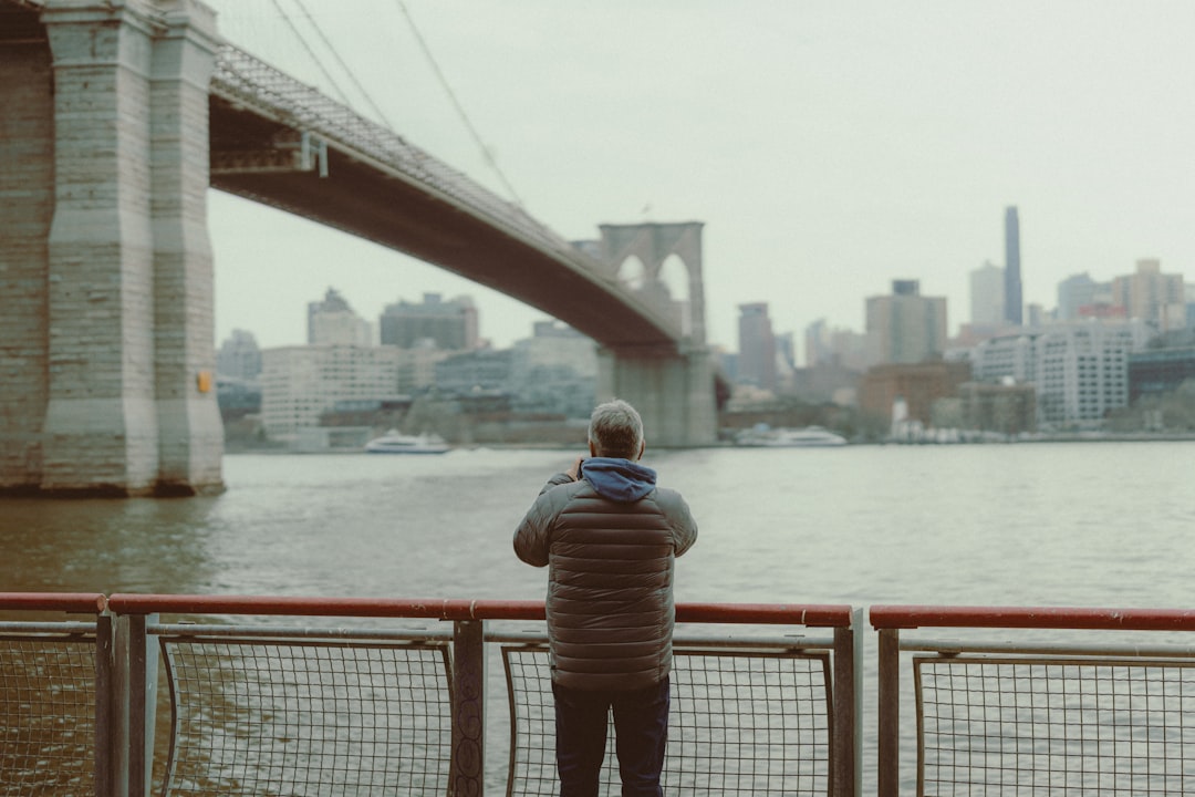 A man stands at a railing, gazing out at the water with a bridge and cityscape in the distance.