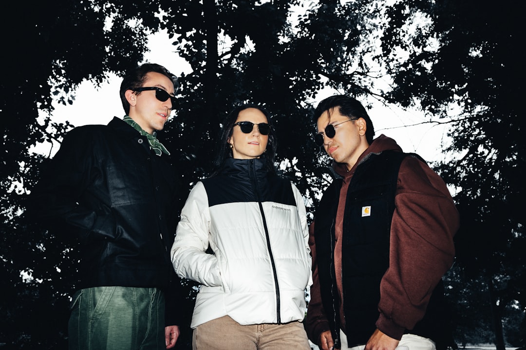 Three individuals stand together, their faces obscured by sunglasses, set against a backdrop of dark trees and a gray sky.