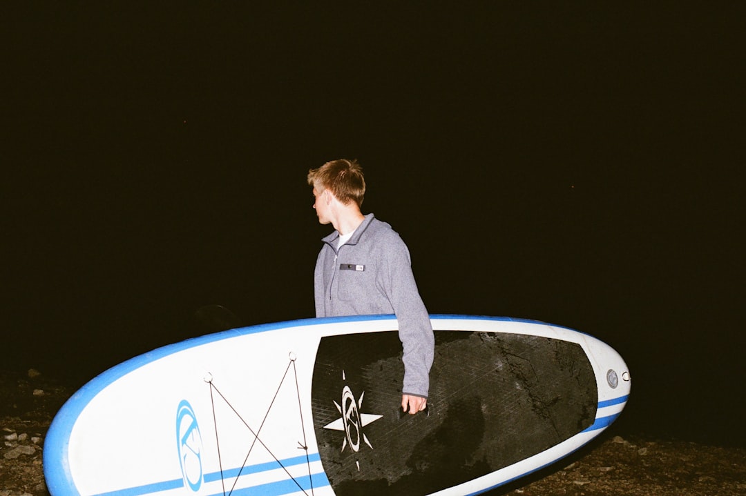 A man stands beside a paddleboard, set against a dark background.