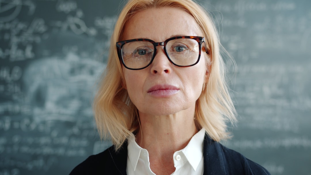 A woman with blonde hair and black glasses stands in front of a chalkboard.