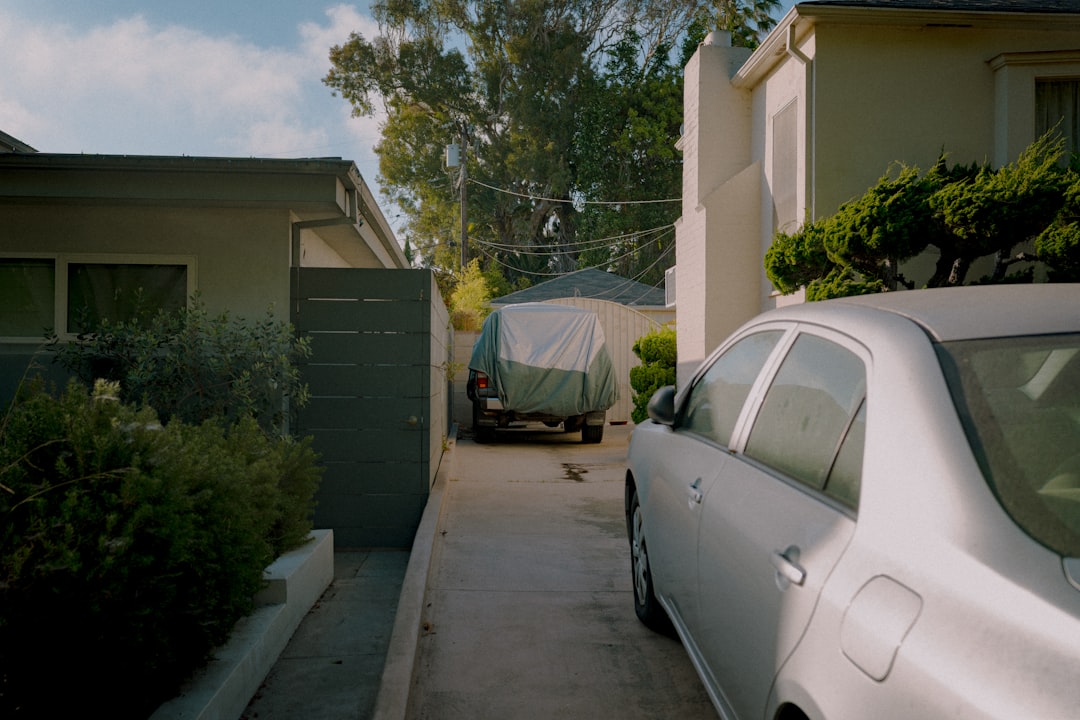 A white car is parked in a narrow driveway, with a covered truck parked behind it.
