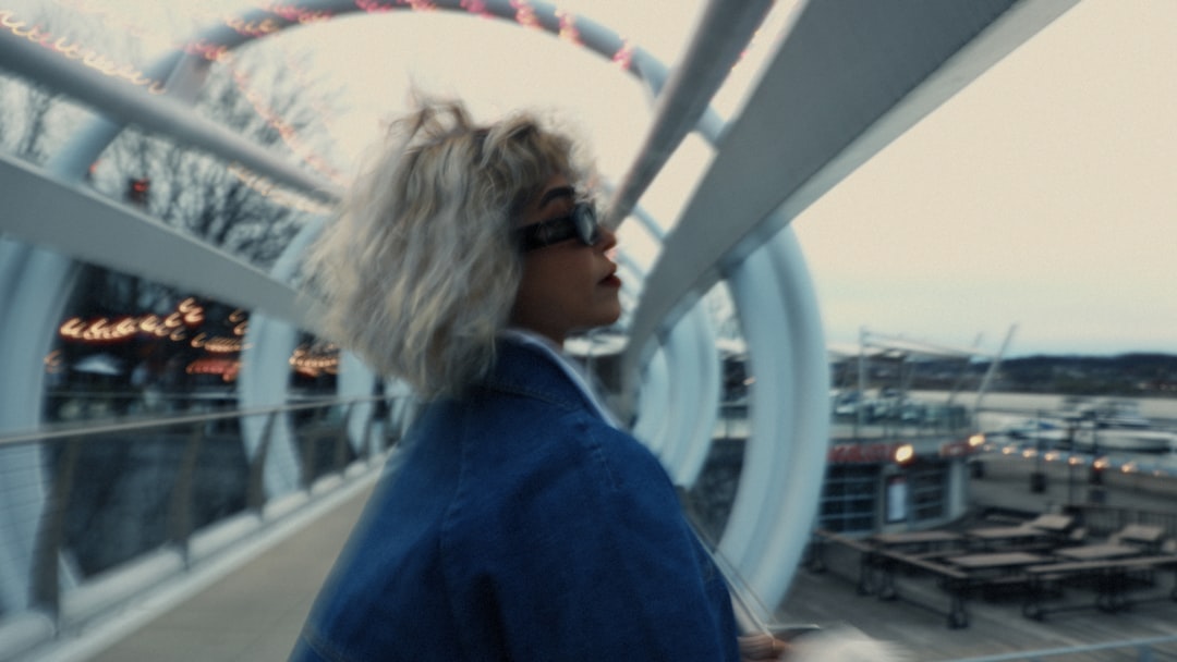 A woman with short, tousled white hair stands on a bridge, gazing to the right.