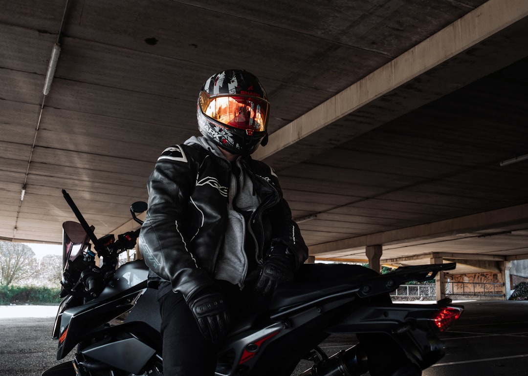 A person sits on a sleek black motorcycle in a concrete parking garage, clad in a black leather jacket and helmet.