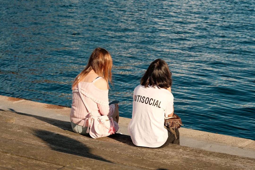 Two women sit on a concrete ledge, facing the water.
