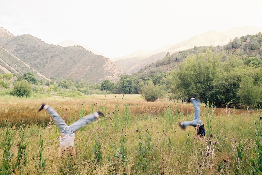 Two individuals perform a handstand in a lush, grassy field surrounded by majestic mountains.