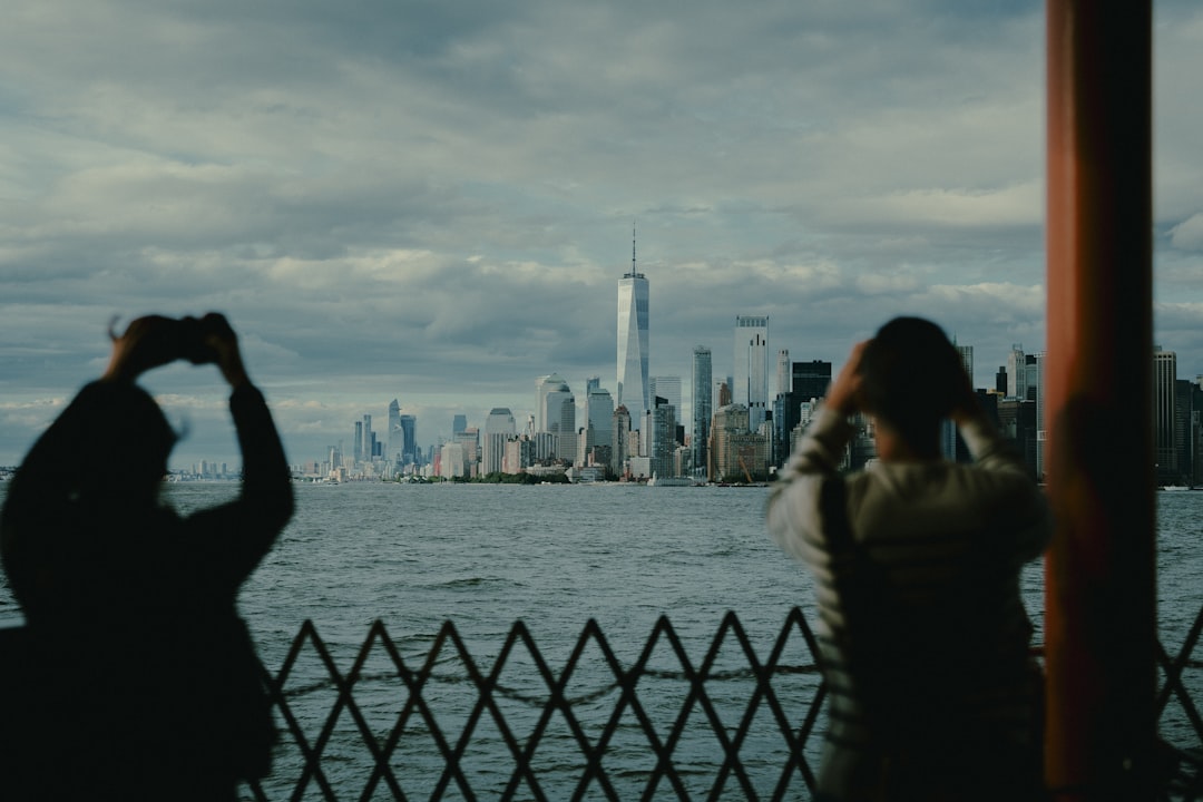 A city skyline is visible across a body of water, with silhouettes of people taking pictures in the foreground.