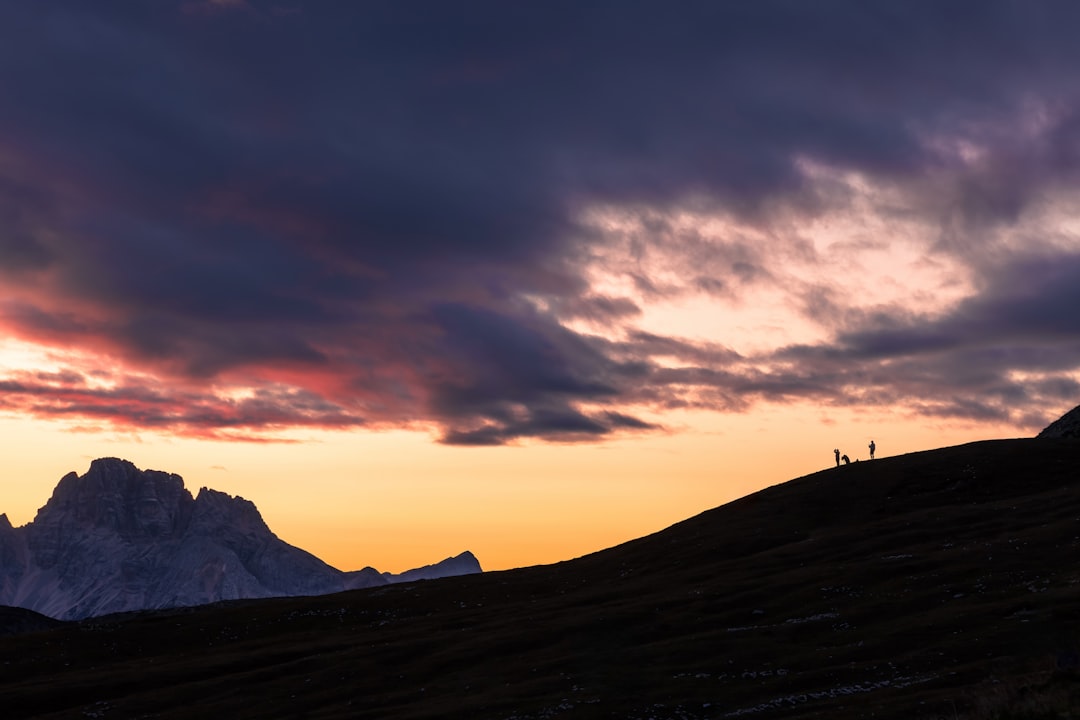 Three silhouetted figures stand atop a dark hill, set against a vibrant orange and purple sky.