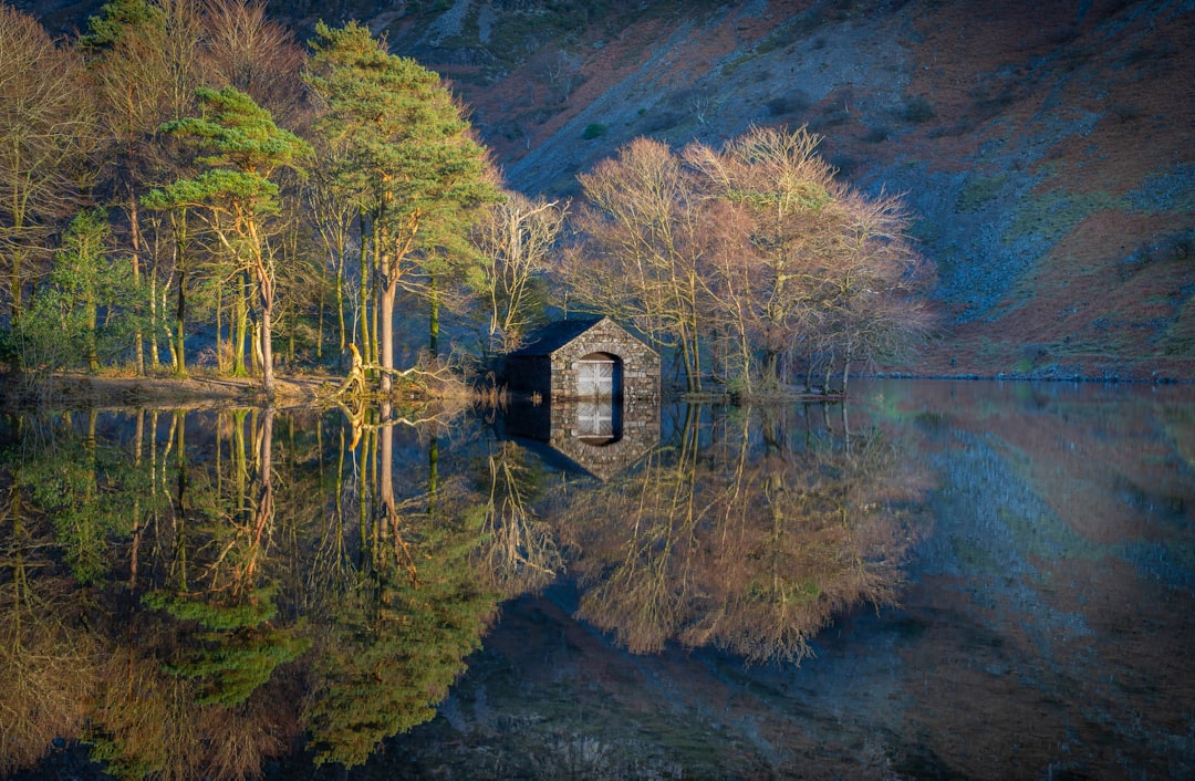 A serene stone building with a large window sits on the edge of a tranquil lake, surrounded by lush trees and reflecting the surrounding landscape.