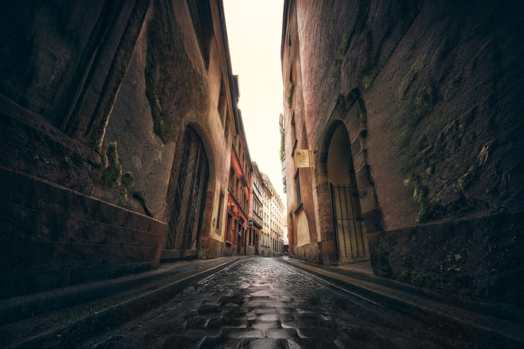 A narrow, cobblestone street flanked by ancient stone buildings, with arched doorways and moss-covered walls.