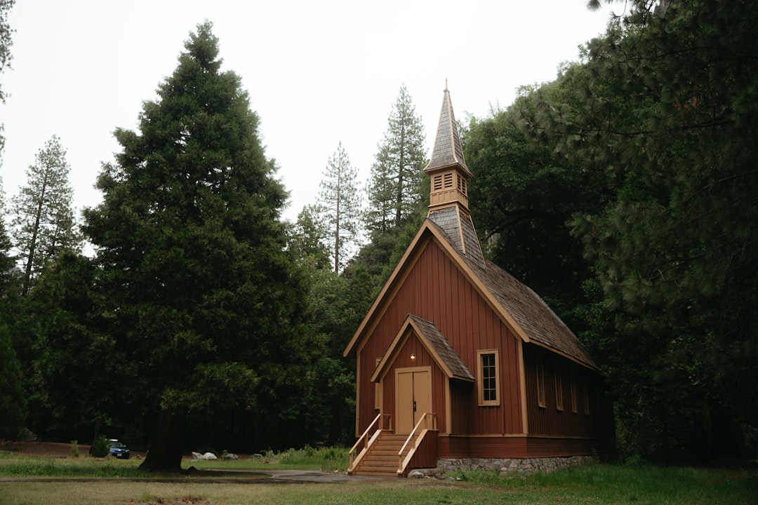 A small, rustic chapel with a steeple stands in front of a lush forest, its wooden exterior and stone foundation blending harmoniously into the natural surroundings.