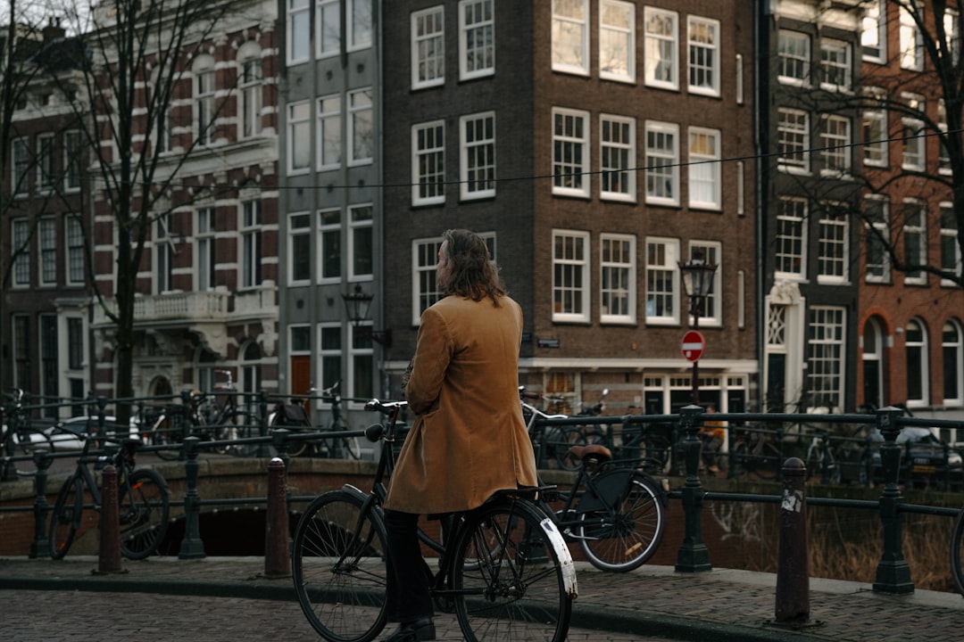 A woman in a tan coat stands with her bicycle in front of a canal and a row of brick buildings.