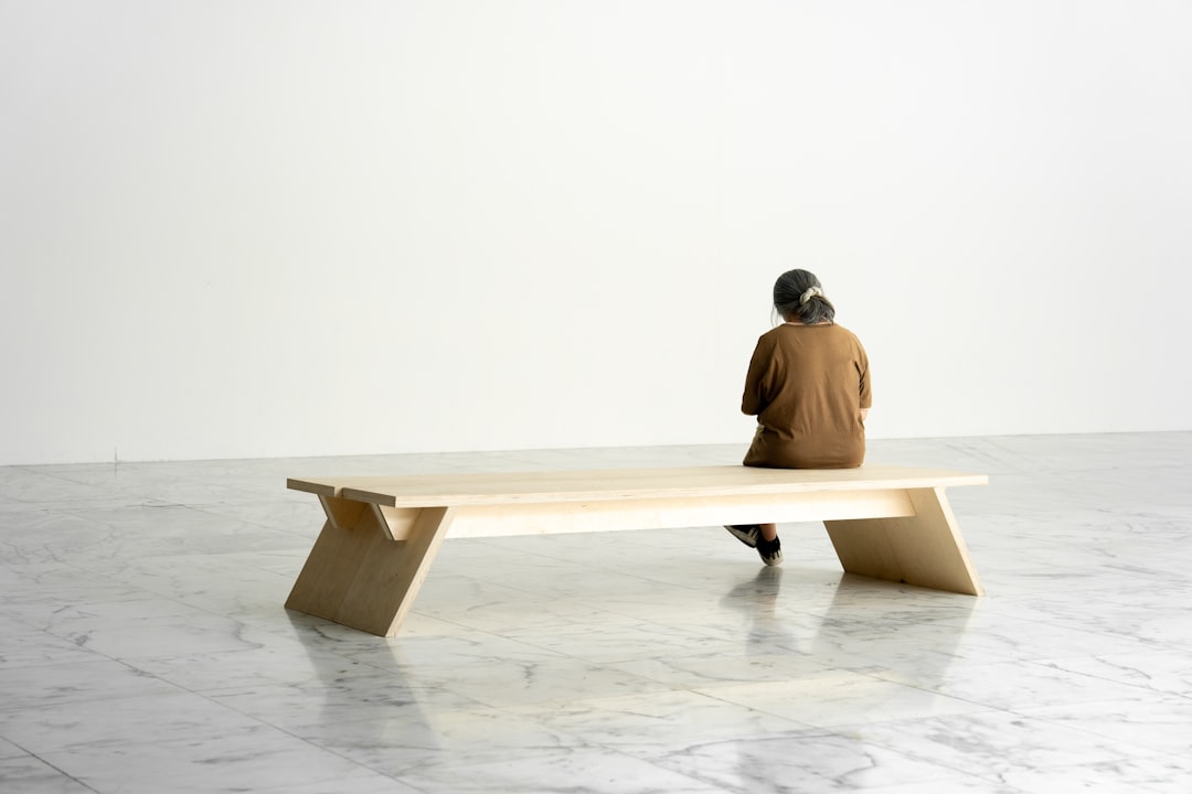 A person sits on a long, light-colored bench in a spacious room with white marble floors and a plain white wall.