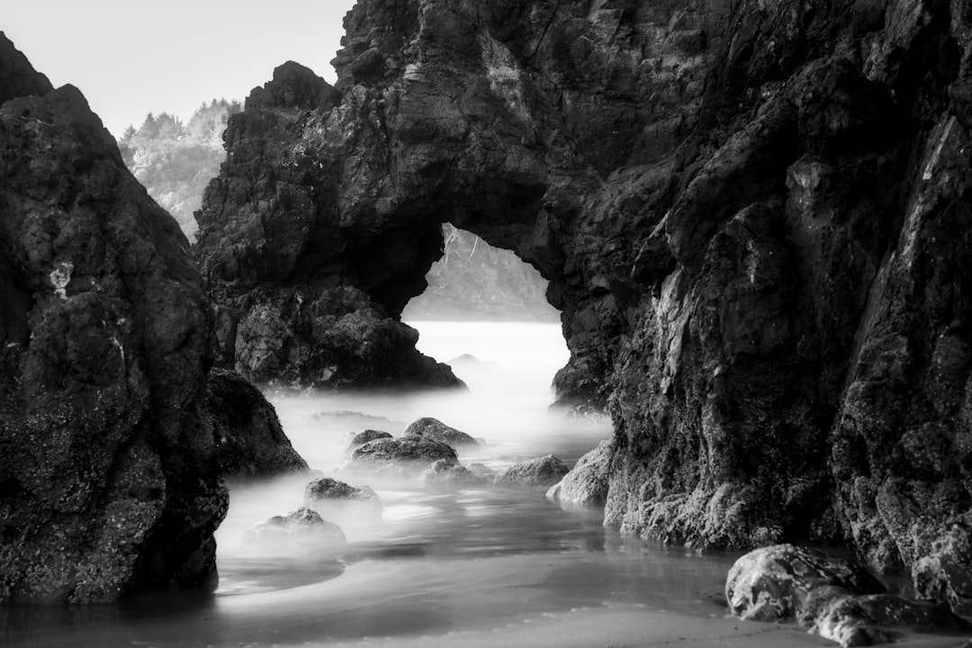 A rugged, rocky archway stands sentinel over a serene beach, its weathered surface bearing the scars of countless tides.