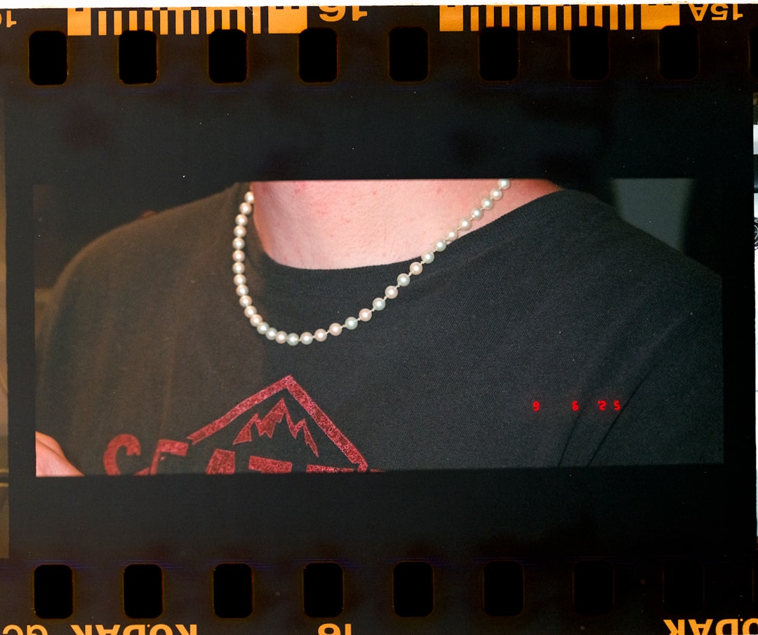 A person wears a pearl necklace over a black shirt with a red logo.
