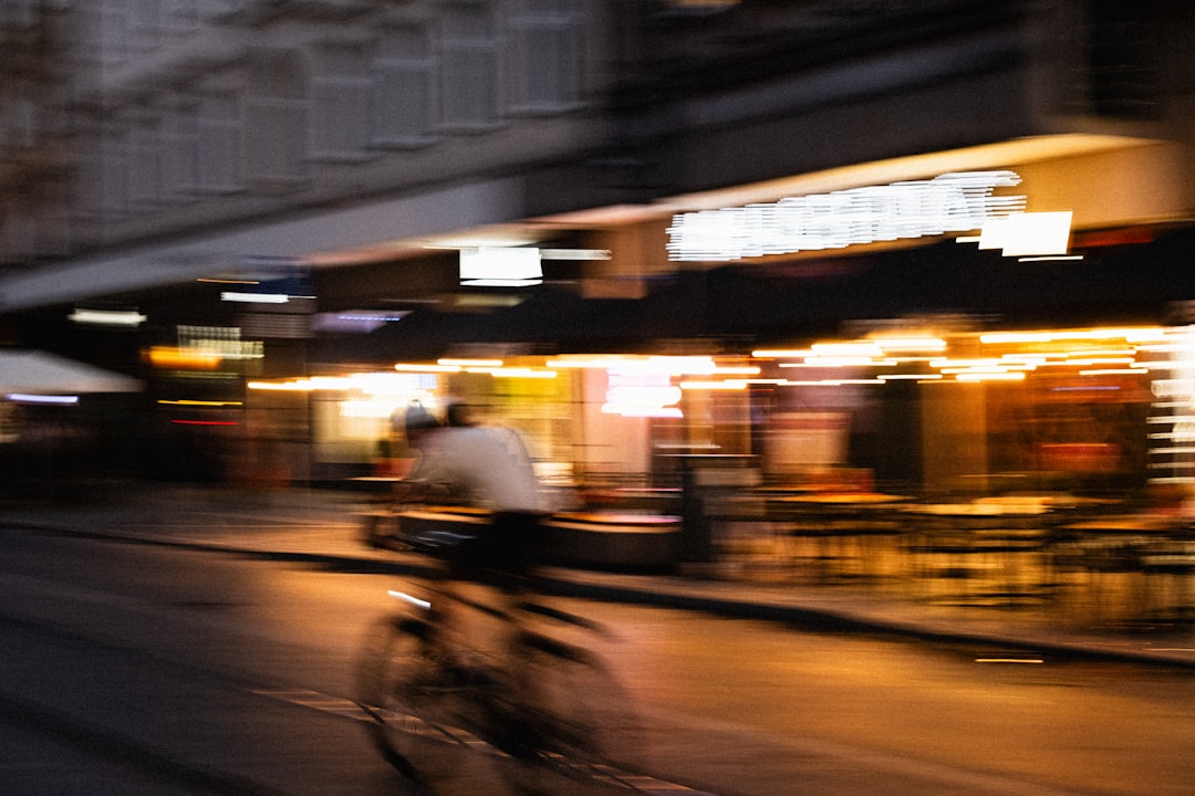 A cyclist pedals past a row of shops, their bright lights reflecting off the wet pavement.