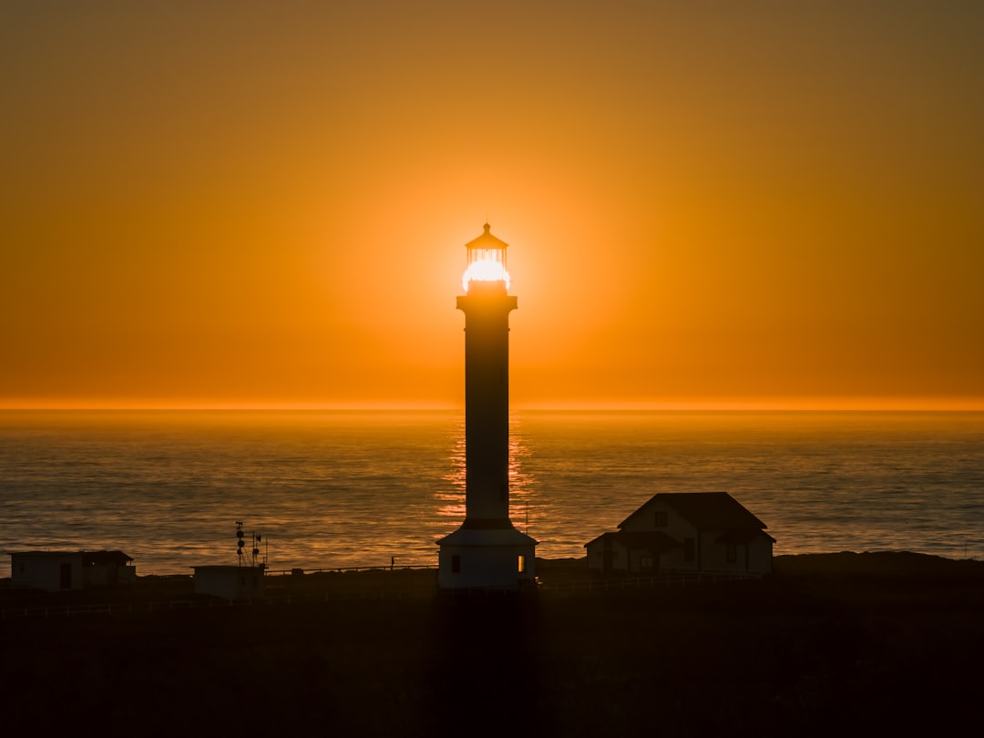 A majestic lighthouse stands tall at sunset, its light shining brightly against the vibrant orange sky.