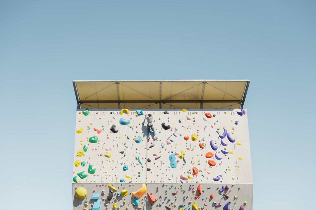 A person climbs a vibrant, multi-colored climbing wall against a clear blue sky.
