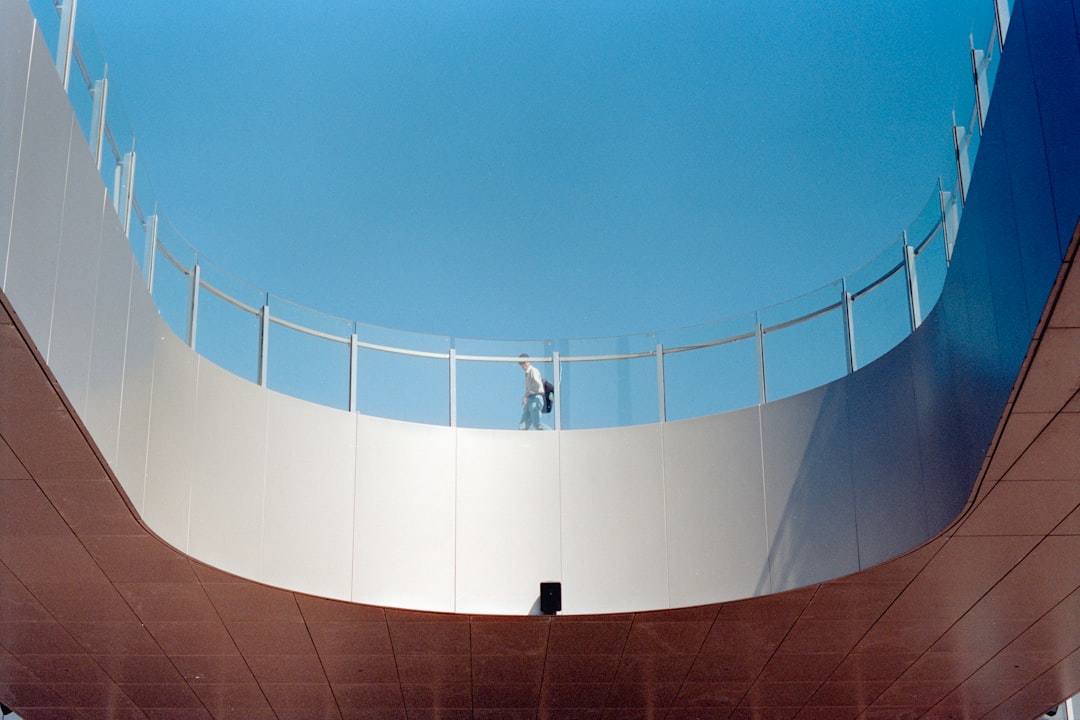 A person stands on a curved, glass-railled balcony, set against a brilliant blue sky.