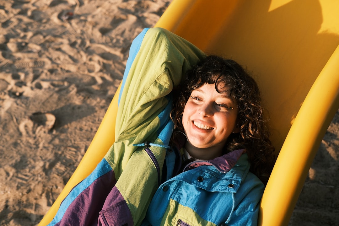 A person with curly brown hair lies on a yellow slide, wearing a jacket with a vibrant color block pattern.