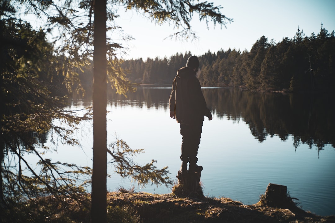 A man stands on a mossy rock, gazing out at a serene lake surrounded by trees.