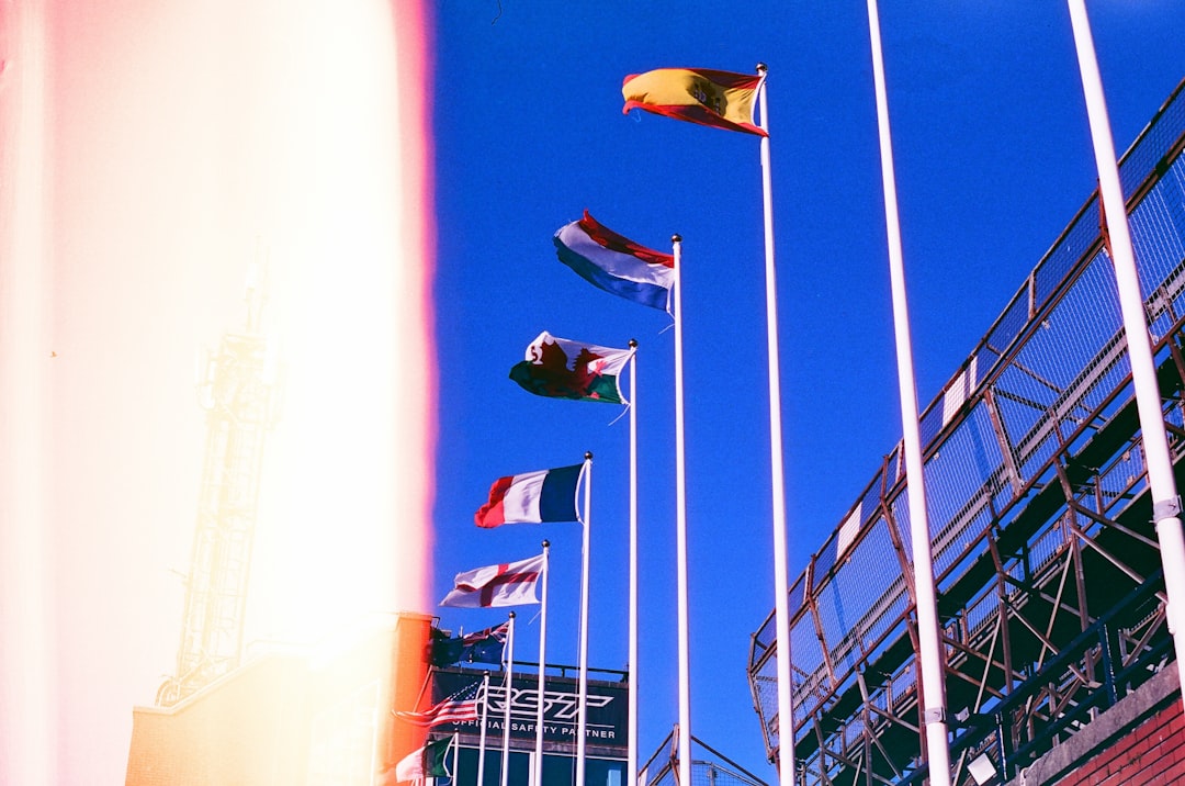 A row of flags flutters in the wind, set against a backdrop of a stadium with bleachers.