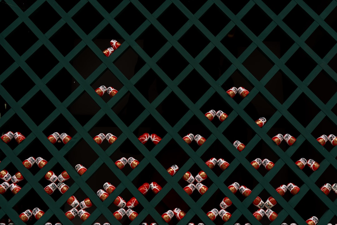 A lattice fence supports rows of small, red, white, and gold objects, likely lanterns, against a black background.