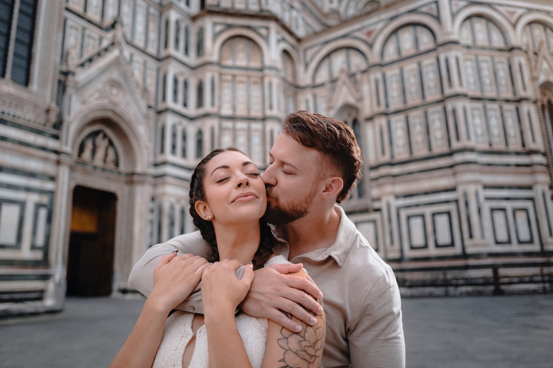 A man with dark hair and a beard stands in front of a large building, his face inches from a woman's face.