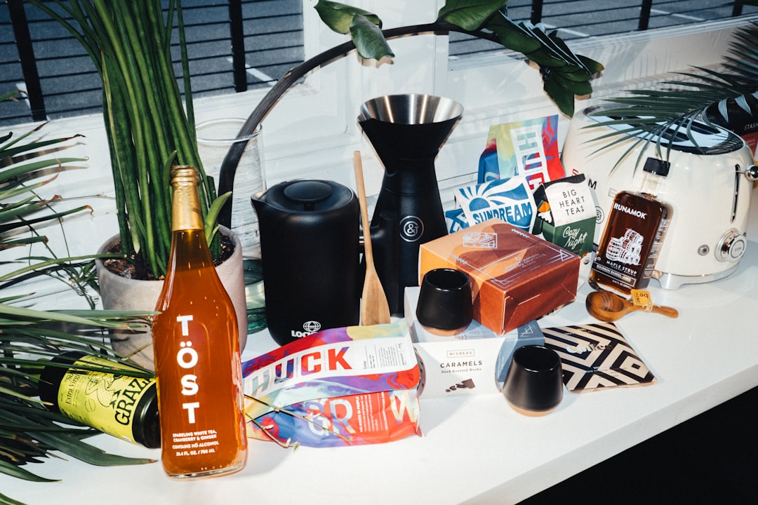 A sleek black coffee grinder sits on a white table, surrounded by an assortment of items, including a bottle of Töst orange-colored liquid, a white toaster, and various other kitchen items.
