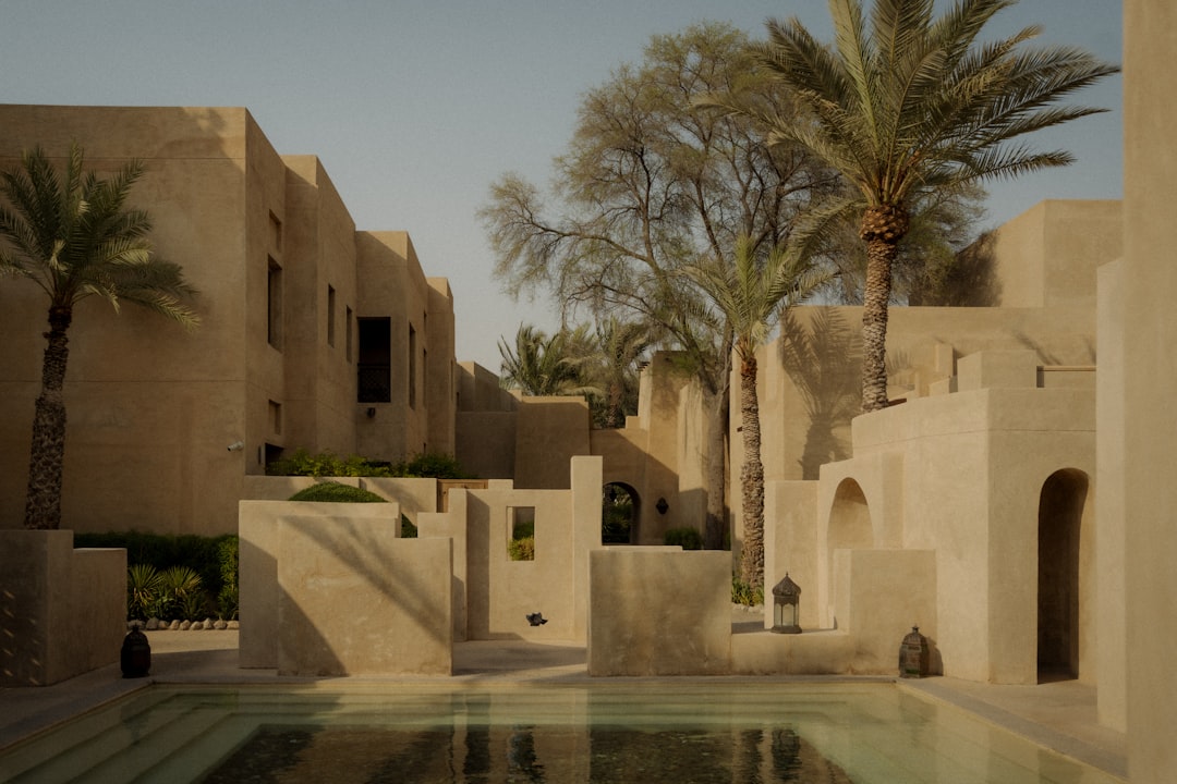 A serene courtyard with a pool and palm trees, surrounded by tan buildings.