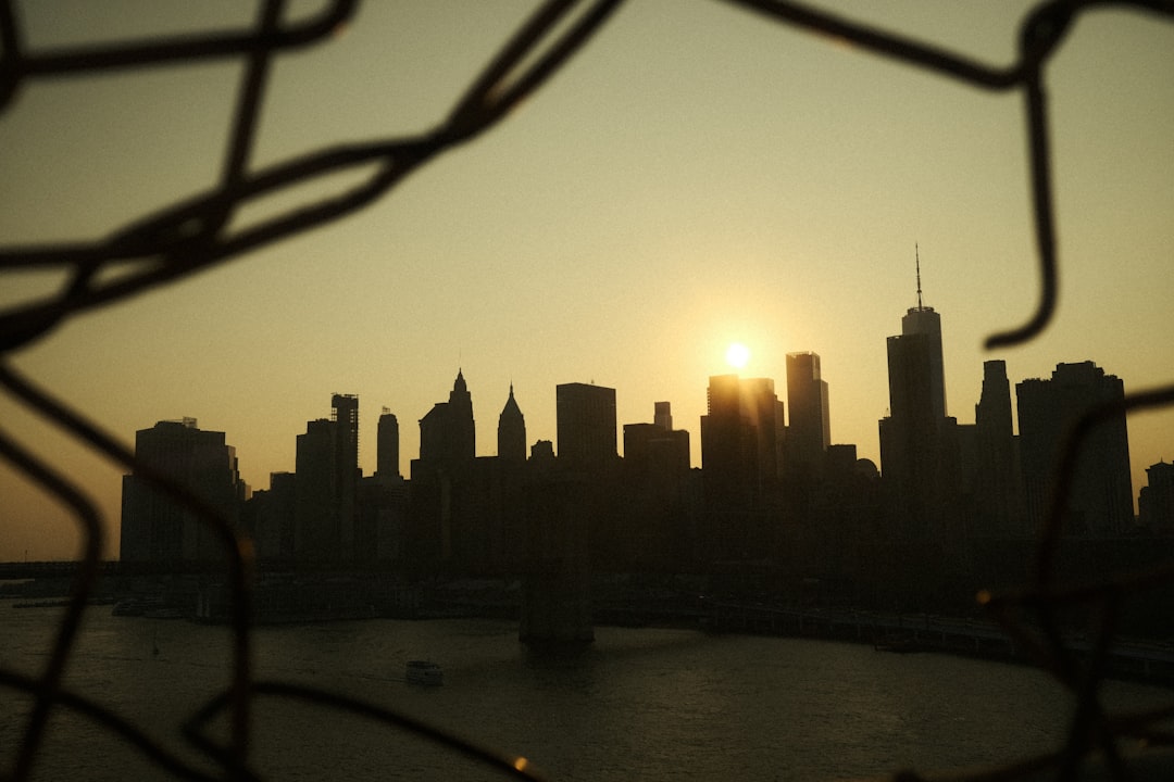 A city skyline silhouetted against the setting sun, viewed through a chain-link fence.