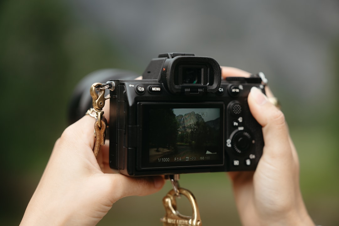 A black camera with a viewfinder and buttons is held by two hands, displaying a blurred image of a rocky mountain on its screen.
