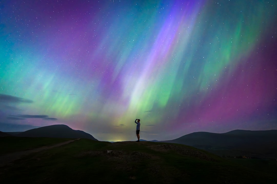 A lone figure stands at the edge of a vast, dark landscape, silhouetted against a breathtaking display of the aurora borealis.