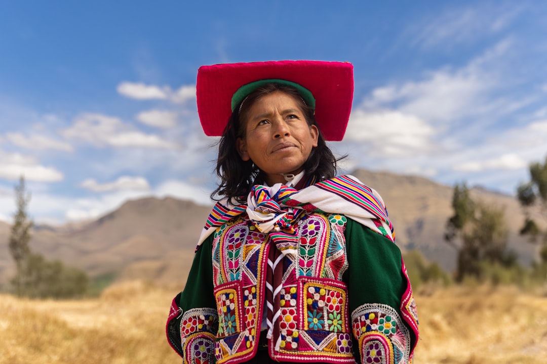 A woman stands in a field, her vibrant green and red embroidered jacket and matching hat a striking contrast to the dry, golden grass beneath her feet.