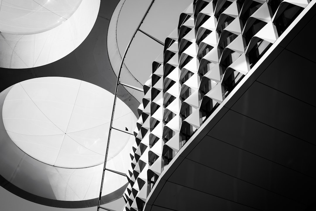 A black-and-white photograph of a staircase with a geometric railing, viewed from the bottom looking up.