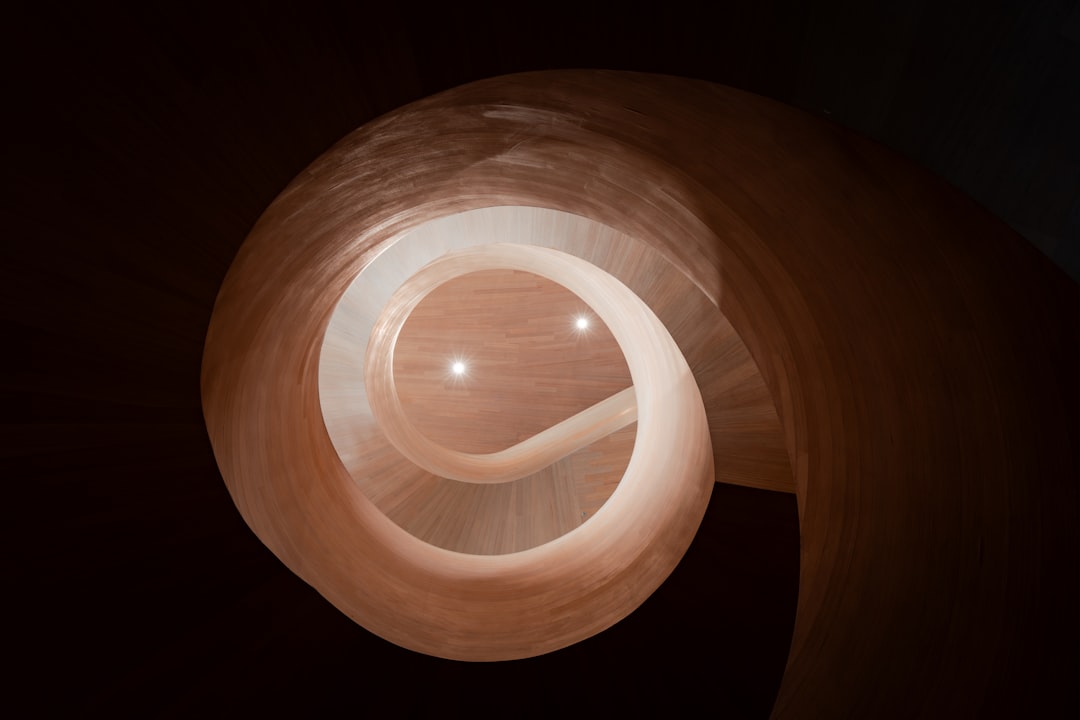 A spiral staircase crafted from light-colored wood dominates the frame, its central axis illuminated by three bright lights, set against a stark black background.