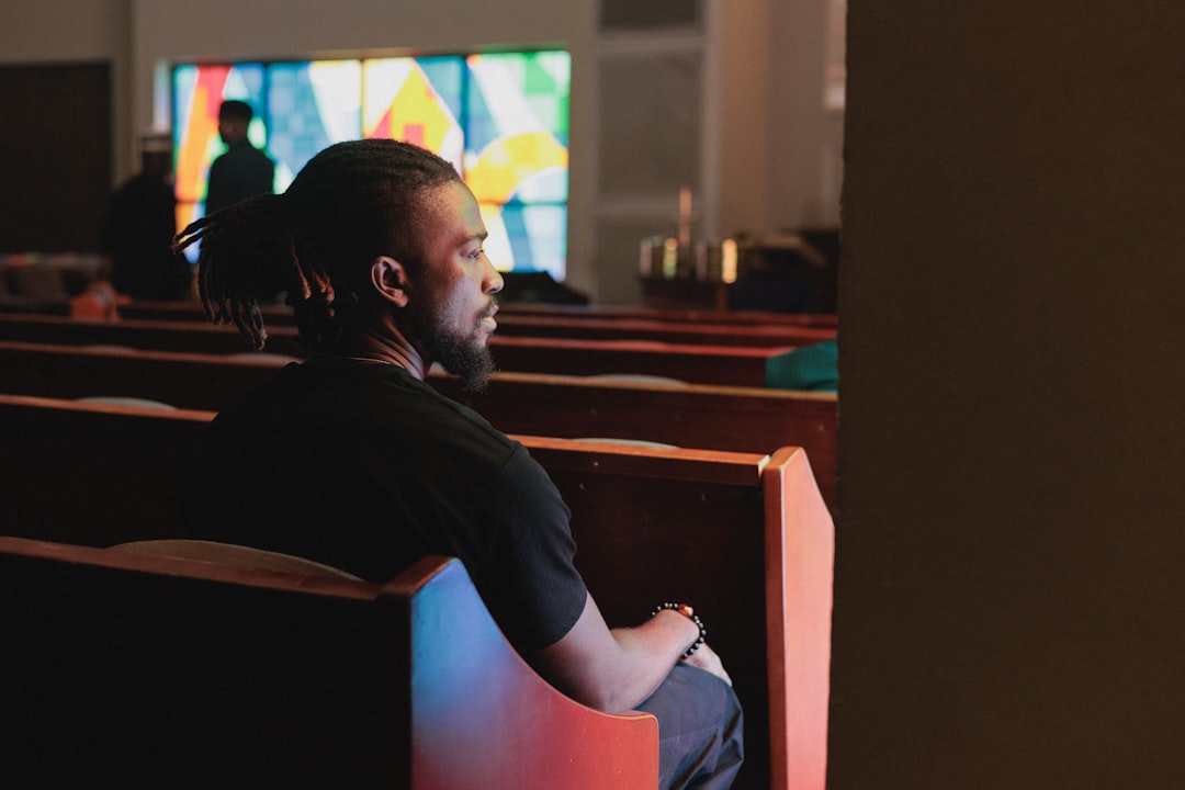 A man with dark skin and black hair sits in a church pew, surrounded by other pews and a stained glass window.