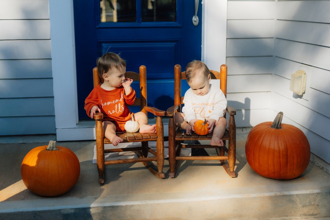 Two babies sit on wooden chairs, surrounded by pumpkins.