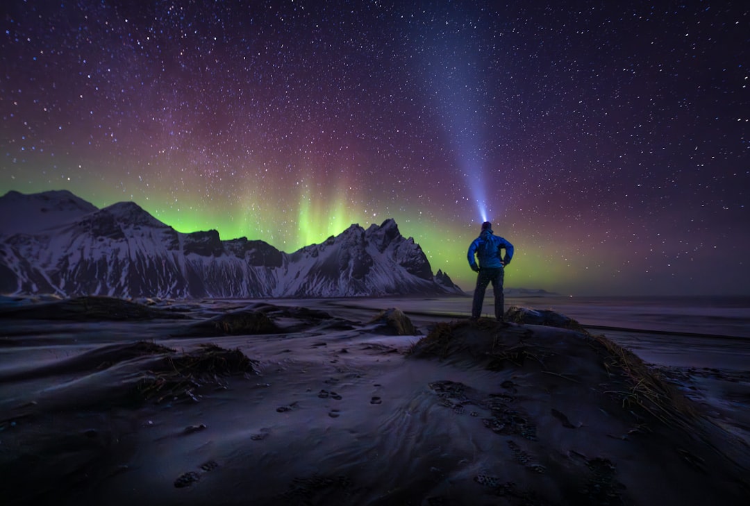 A person stands on a beach, gazing at the Northern Lights dancing across the night sky.