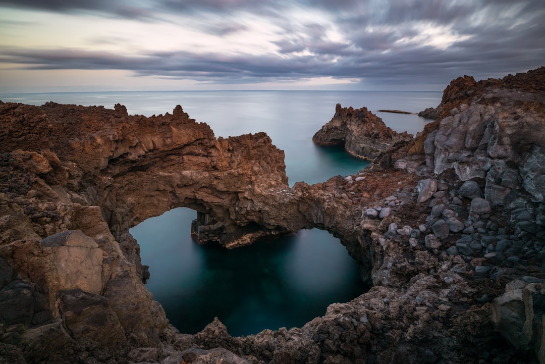 A natural archway stands amidst rugged, rocky terrain, with a serene body of water flowing through its center.