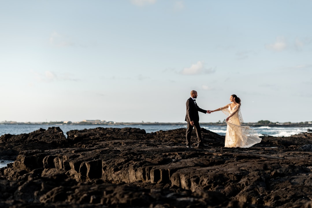 A bride and groom hold hands on a rocky shore, with the bride's white dress and veil fluttering in the wind.