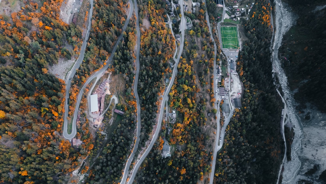 A winding road traverses a mountainous terrain, flanked by vibrant trees and a soccer field.