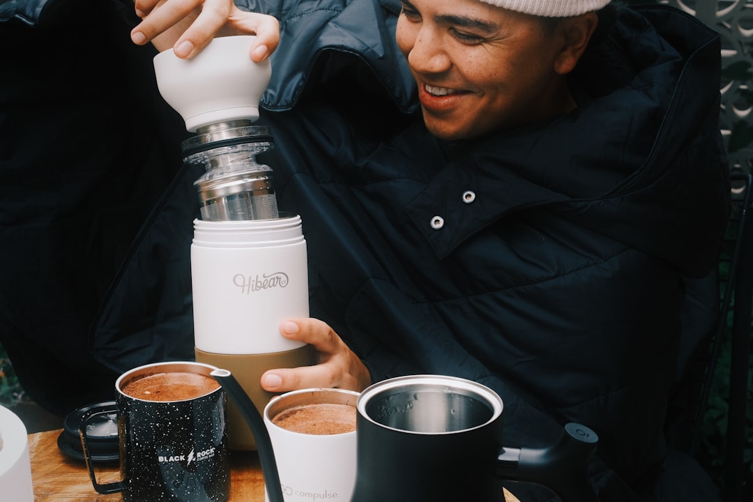 A man in a black coat and white hat is pouring coffee from a white container into a cup.