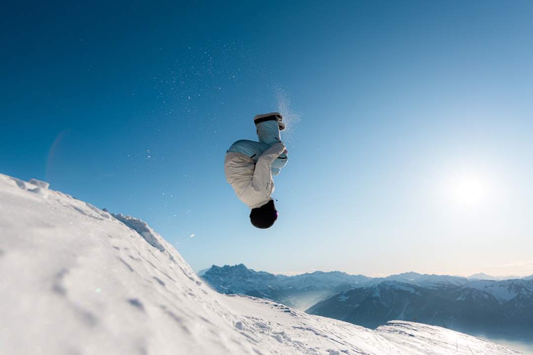 A person in a white coat and black hat executes a backflip in mid-air, set against a snowy mountainous backdrop.