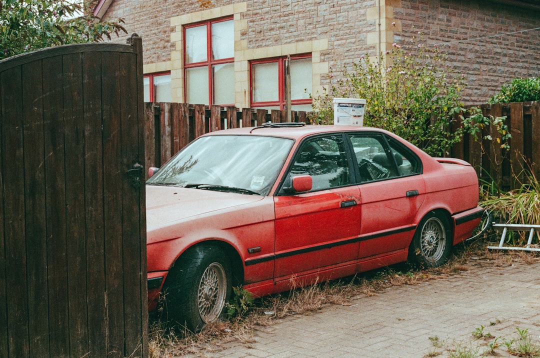 A red car sits in front of a wooden fence, its faded paint and dusty appearance suggesting a state of neglect.