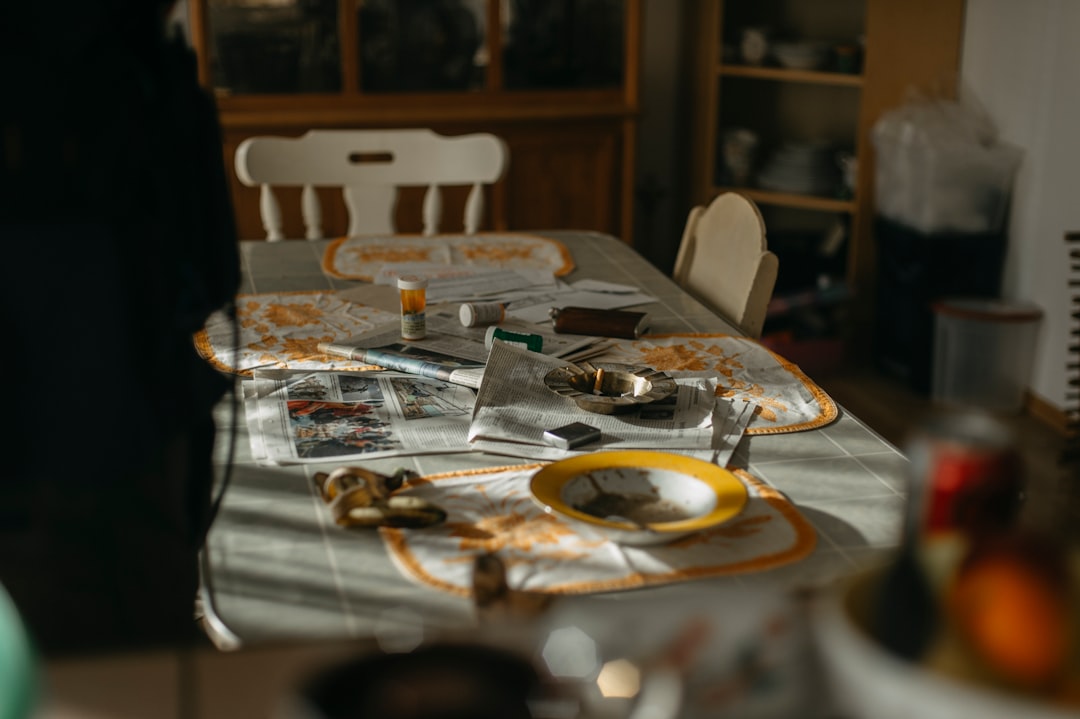 A table with a yellow and white tablecloth, adorned with various items, including a yellow plate, newspapers, and a pill bottle, in a dimly lit room.